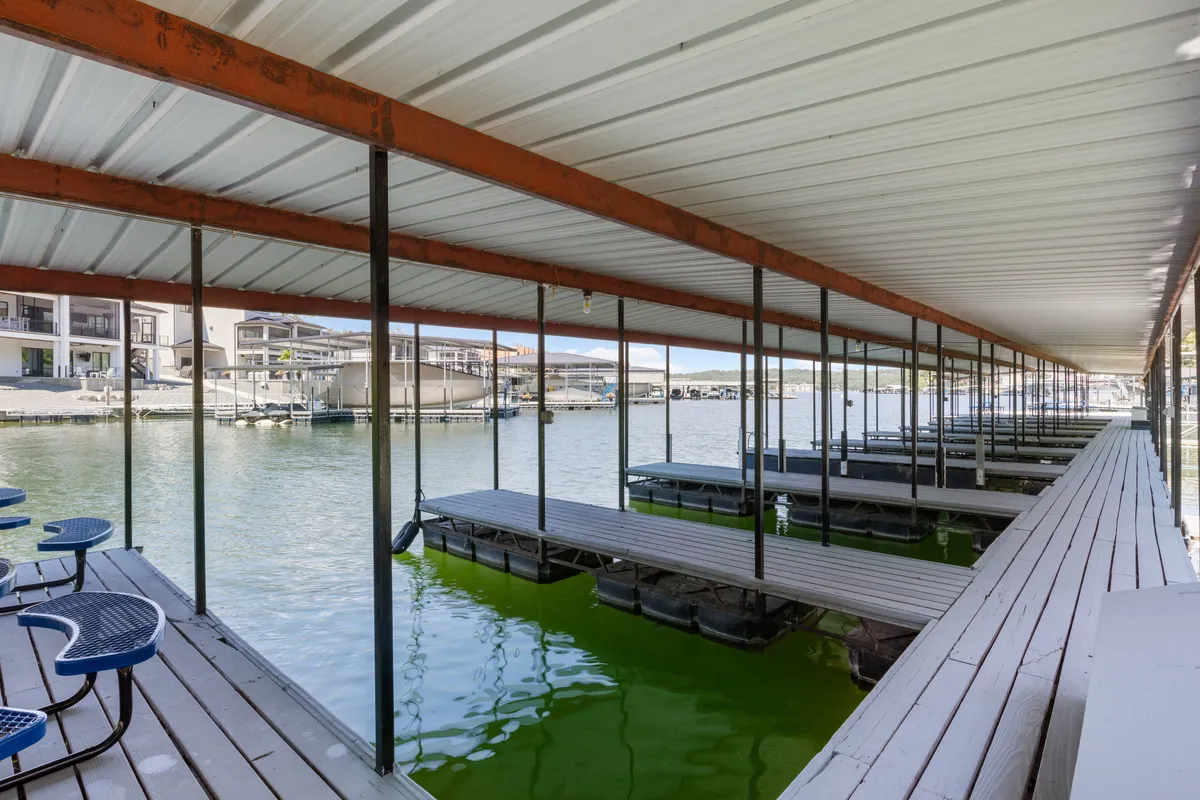 A calm lakeside dock stretches over the water, with boats moored nearby and the tranquil water surface reflecting the sky. The scene is open, inviting guests to walk along the dock or relax at its edge. Grassy areas and trees can be glimpsed in the background, offering a natural, pet-friendly environment.