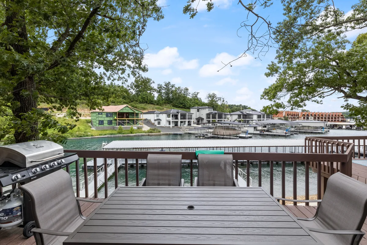 An outdoor dining set and grill are positioned by the water, with distant houses across the lake. The setting creates a cozy, intimate spot for sunset dinners or unwinding together, with a relaxed lakeside feel.