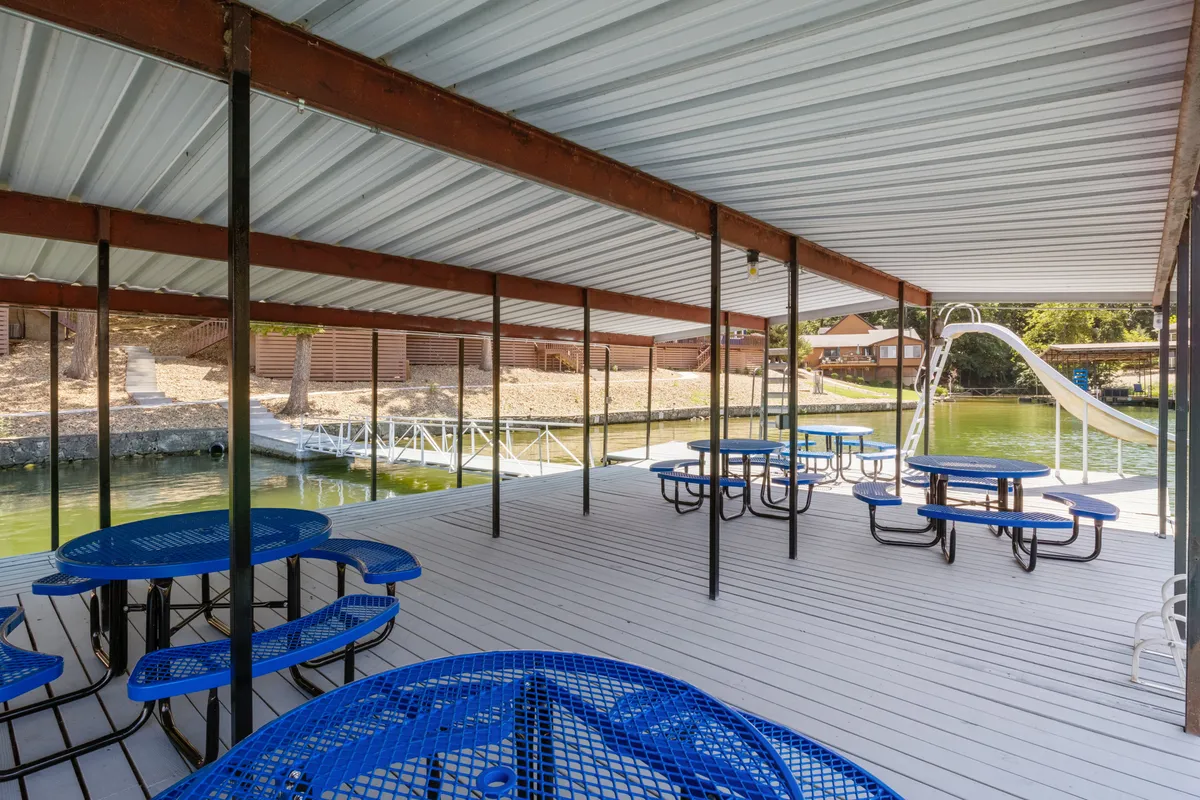 This image showcases a vibrant lakeside dock area featuring bright blue picnic tables, a water slide extending into the lake, calm waters, and a lush surrounding landscape. The inviting outdoor space is clearly designed for communal enjoyment, water activities, and relaxation.