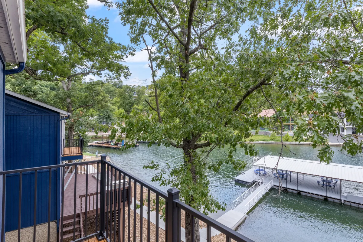 A tranquil lakeside view showing the dock extending over calm water, with trees framing the scene and a balcony railing in the foreground. The setting sun casts a soft glow across the lake, creating a peaceful and romantic atmosphere ideal for couples seeking relaxation.