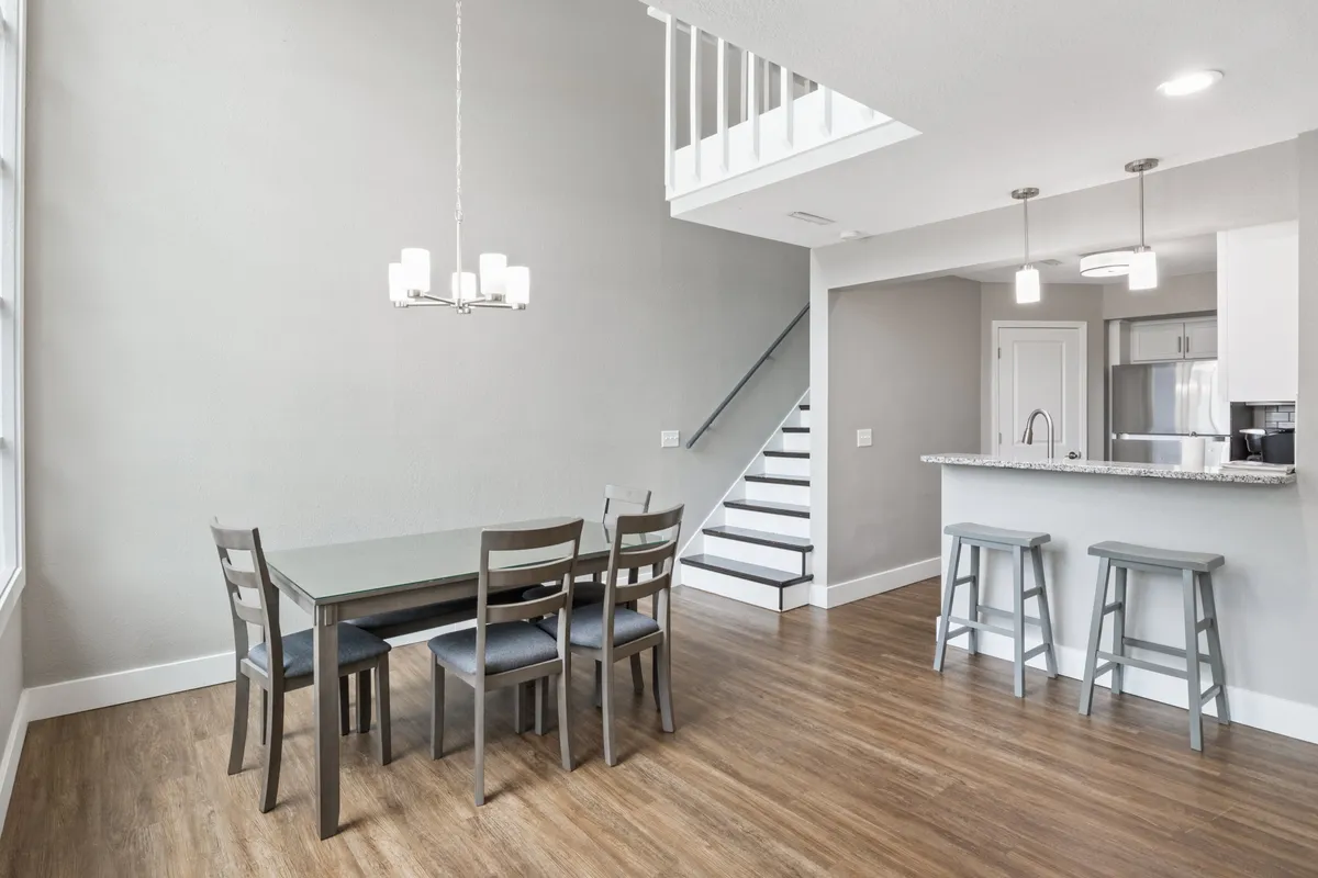 A contemporary dining area is shown, featuring a stylish dining table beneath a chandelier, with a staircase and kitchen bar in view. The setting is modern and cozy, perfect for family meals or entertaining guests.