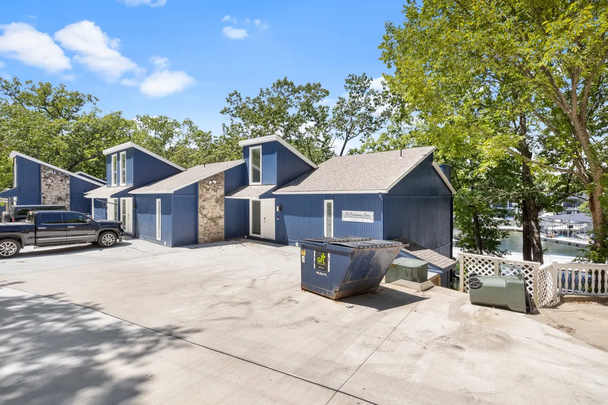 The exterior of a blue house framed by stone accents and surrounded by mature trees, with a generous driveway and hints of open green space. The setting feels relaxed and welcoming, providing plenty of room to explore or rest outside.