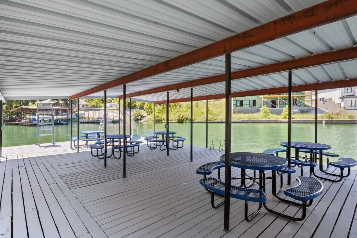 A casual waterfront gathering area featuring picnic tables under a metal roof, with a clear view of the water just steps away. The relaxed setup is ideal for parents to watch over their children or enjoy a sunny afternoon by the lake.